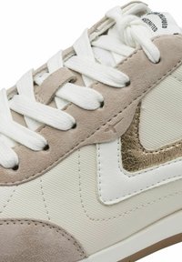 Sneaker beige e bianco con materiali in suede e sintetico testurizzato, dotato di lacci bianchi, un accento metallico dorato e dettagli cuciti.