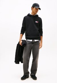 Čierna mikina s kapucňou s nášivkou s logom, sivé džínsy a čierne topánky. Drží čiernu bundu a zrolovaný uterák. Doplnok: čierna šiltovka.