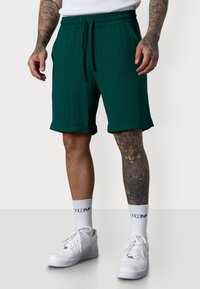 Man wears green textured shorts, white socks, and sneakers, showcasing intricate leg tattoos, with a neutral background.