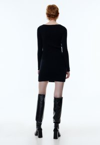 Schwarzes, geripptes Langarmkleid mit taillierter Silhouette, verfügt über einen Rundrücken und einen hohen Schlitz. Kombiniert mit knielangen schwarzen Lederstiefeln.