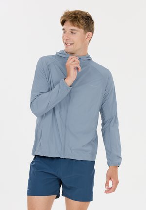 Junger Mann lächelt, trägt eine hellblaue Kapuzenjacke und dunkelblaue Shorts, zieht mit der rechten Hand am Reißverschluss der Jacke.