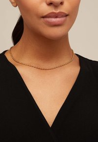 Collar de cadena en tono dorado con eslabones redondeados y alargados. Lleva puesto alrededor del cuello, complementando una prenda negra con escote en V.