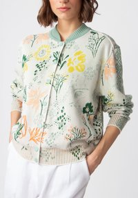 Stickad cardigan med ett blommönster i grönt, gult och orange på en ljus bakgrund, med ribbad mudd och knappstängning.