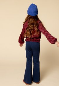 Rode bloempatron blouse met ballonmouwen, gecombineerd met marineblauwe flared broek. Het model draagt een blauwe gebreide beanie. De achtergrond is lichtbeige.