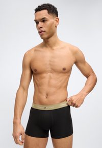 Άνδρας χωρίς πουκάμισο διορθώνει τη μέση των μαύρων μποξεράδικων Tommy Hilfiger, κοιτάζοντας προς τα αριστερά του μπροστά σε έναν απλό φωτεινό φόντο.