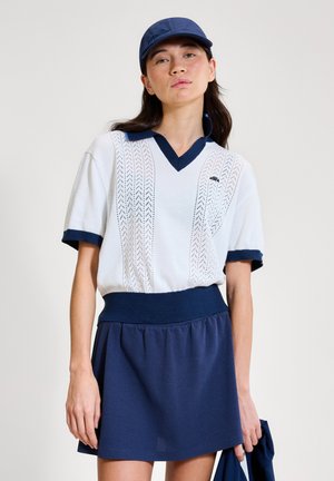 Woman wearing white knit polo shirt with navy trim, navy skirt, and navy cap, holding matching navy garment in right hand.