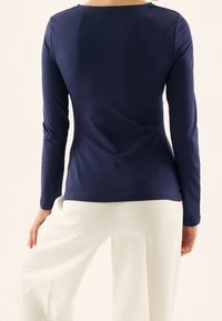 Marinblå långärmad topp i slätt tyg, med rundad halsringning och figurnära silhuett, som bärs tillsammans med vita vida byxor.