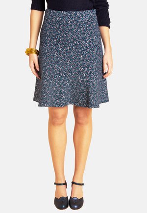 Frau trägt einen blau gemusterten knielangen Rock, schwarze Schuhe mit Knöchelriemen, schwarzes Oberteil und goldene Armreifen am linken Handgelenk.