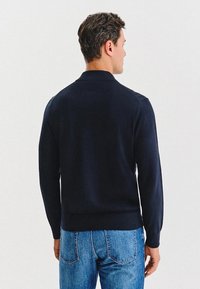 Mężczyzna odwrócony plecami, ubrany w ciemnogranatowy sweter z długim rękawem i niebieskie dżinsy, na jasnym, jednolitym tle.