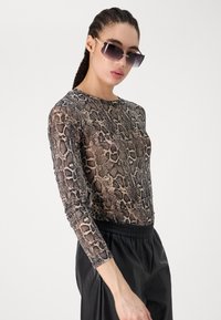 Transparante top met lange mouwen in een bruin en zwart slangenschubbenpatroon, gecombineerd met zwarte broek met elastische taille. Model draagt een donkere zonnebril.