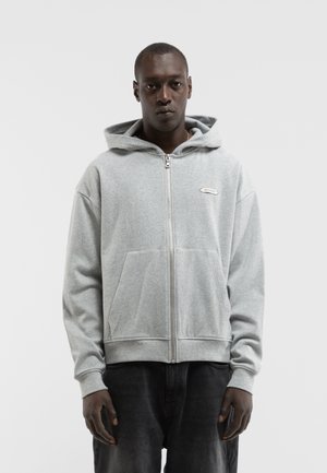 Mann trägt einen hellgrauen Kapuzenpullover mit Reißverschluss und Fronttaschen sowie schwarze Hose, steht vor einem einfachen hellen Hintergrund.