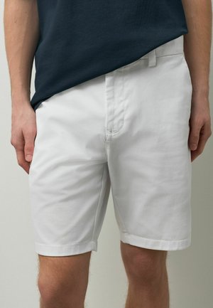 Mann trägt weiße knielange Shorts und ein dunkelblaues T-Shirt, steht mit entspannten Händen seitlich an neutralem Hintergrund.