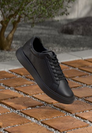 Czarny skórzany sneaker z tłoczonym wzorem i sznurówkami, unoszący się nad drewnianymi płytkami z małymi białymi kamykami pomiędzy nimi na zewnątrz.