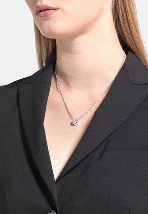 Collar de cadena de plata con un colgante redondo que presenta una piedra clara, llevado sobre un blazer negro, resaltando un diseño minimalista.