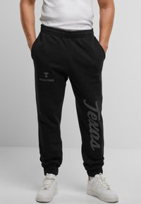 Sorte sweatpants med elastisk talje, med "T" og "UPSCALE STUDIOS" logoer i grå, og "Texas" trykt ned langs siden.