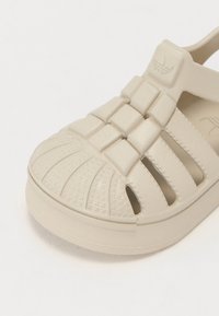 Beige slip-on sandal made of textured rubber, featuring a crisscross design, open sides, and a striped tread on the sole.