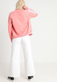 Rosa zip-up jacka med vit kant och tre vita ränder på ärmarna, tillsammans med vida vita byxor och svarta skor.