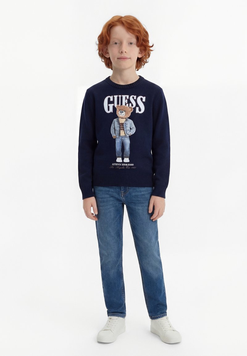 Rudovlasý chlapec stojí, oblečený v námornícko modrom svetri s obrázkom medveďa a nápisom „GUESS“, modrých džínsoch a bielych teniskách na bielom pozadí.