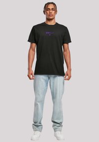 Black cotton t-shirt featuring purple text. The design has a classic crew neck and short sleeves, paired with light blue jeans and white sneakers.