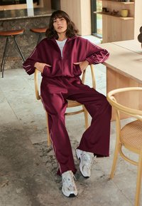 Bordeaux tracksuit met witte zijkanten, bestaande uit een ritsjack en losse broek. Draagt het met witte sneakers, zittend op een houten stoel.