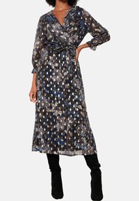 Žena v černých květinových midi šatech s průhlednými dlouhými rukávy a opaskem v pase, v černých kozačkách po kolena, stojící s jednou rukou na bocích.