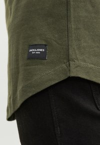 Olivgrön T-shirt med rundad fåll, med en svart "JACK & JONES"-etikett. Texturerad bomullstyg, kombinerat med svarta byxor.