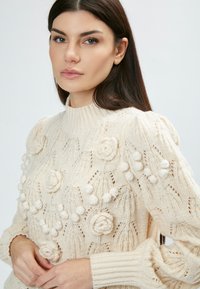 Gestrickter Pullover in Creme mit floralem Muster, Bubbel-Akzenten und hohem Kragen. Texturiertes, dickes Garn mit gerippten Bündchen.