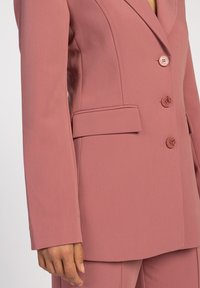 Lichtroze op maat gemaakte blazer met een notch-kraag, drie knopen en zijzakken. Glad oppervlak met subtiele stiksels.
