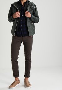 Henkilö säätää tummanvihreää takkia laivastonsinisten ruudullisten paitan päällä, pukeutuneena ruskeisiin housuihin ja vaaleanruskeisiin lenkkareihin, seisoo vaalealla yksinkertaisella taustalla.