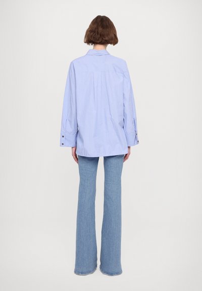 Camicia a righe blu con vestibilità comoda e maniche lunghe, caratterizzata da polsini con bottone e una piega sul retro. Abbinata a jeans svasati azzurri.