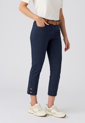 Femme portant un pantalon court bleu marine avec détail de boutons, ceinture marron, haut blanc à manches courtes et baskets blanches, debout la main dans la poche.