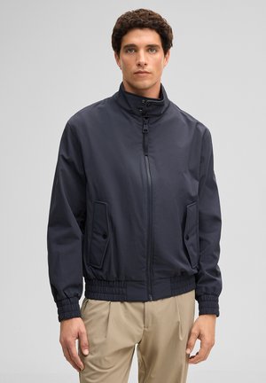 Jeune homme portant une veste zippée bleu marine avec des poches à boutons et un pantalon beige, se tenant devant un fond gris clair uni.