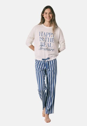 Smilende kvinde iført hvid langærmet top med teksten "Happy is the real treasure" og blå-hvide vertikale stribede pyjamasbukser, barfodet.