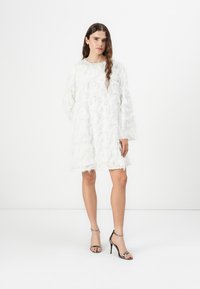 Robe blanche avec des motifs de volants texturés et superposés, à manches longues et coupe ample. L'ourlet effiloché ajoute une touche de détail. Associée à des escarpins métalliques.