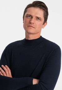 Marineblauer Rundhals-Pullover, stricktexturiert, schlanker Schnitt, lange Ärmel, gerippte Bündchen, getragen von einer Person mit verschränkten Armen vor einem weißen Hintergrund.