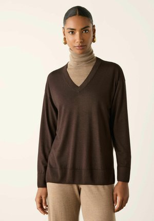 Kvinde iført en brun V-hals trøje over en beige rullekrave og beige bukser, stående mod en ensfarvet off-white baggrund.