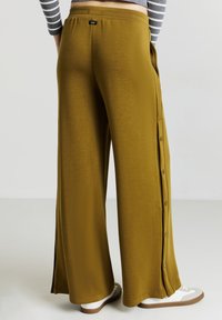 Pantalons larges vert olive avec une texture lisse, des détails de boutons sur le côté et une coupe décontractée. Portés avec des baskets blanches et grises.