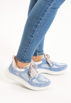 Person wearing light blue suede and mesh sneakers with beige laces and dark blue skinny jeans, standing on a white surface.