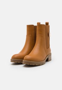 Bottines en cuir tan avec un petit talon empilé et une semelle texturée, mettant en avant des panneaux latéraux élastiques et un détail de sangle décorative.