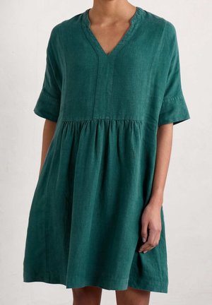 Frau trägt ein locker sitzendes, knielanges grünes Kleid mit kurzen Ärmeln und V-Ausschnitt und steht vor einem schlichten hellen Hintergrund.