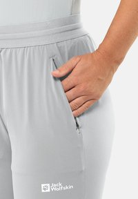 Lichtgrijze, lichte broek met een gladde textuur, voorzien van een ritszak aan de zijkant en een elastische tailleband. Jack Wolfskin-logo zichtbaar.