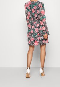 Lyhythihainen kukkamekko, jossa on musta tausta, vaaleanpunaisia ja vihreitä kukkakuoseja sekä röyhelöhelma, yhdistettynä valkoisiin avokkaisiin.