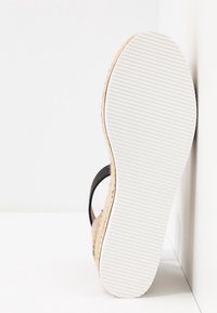 Semelle blanche texturée d'une sandale compensée avec une plateforme en jute beige tressée et une bride noire, positionnée à la verticale contre un angle blanc.