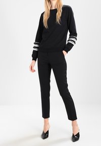 Svart sweatshirt med vita randiga detaljer på ärmarna, tillsammans med skräddarsydda svarta byxor och svarta spetsiga klackskor. Slät textur.