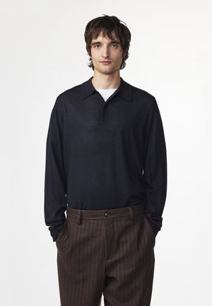 Młody mężczyzna z ciemnymi włosami nosi czarną koszulkę polo z długim rękawem na białej koszulce i brązowe spodnie w cienkie prążki, stoi z rękami w kieszeniach.