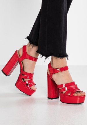 Escarpins en cuir verni rouge avec des talons blocs épais, des brides croisées et un détail de boucle à la cheville. Jeans noirs effilochés visibles.