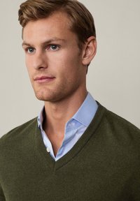 V-neck, olive green sweater layered over a light blue collared shirt. Smooth texture, fitted design, and subtle ribbed detailing at edges.