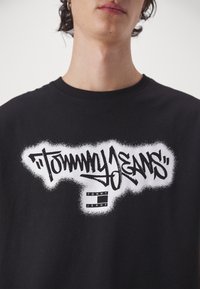 Black cotton t-shirt featuring a large white graffiti-style logo saying "TOMMY JEANS," with a rectangular label underneath, textured print.