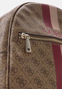 Sac à dos marron à motifs avec une quincaillerie dorée, comportant une poche zippée, des accents rayés bordeaux et beige, et un détail de logo en relief.