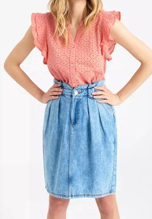 Mujer que lleva una blusa de calados de manga corta de color coral rosa metida en una falda vaquera azul claro de talle alto con pliegues y una cinturilla ancha.
